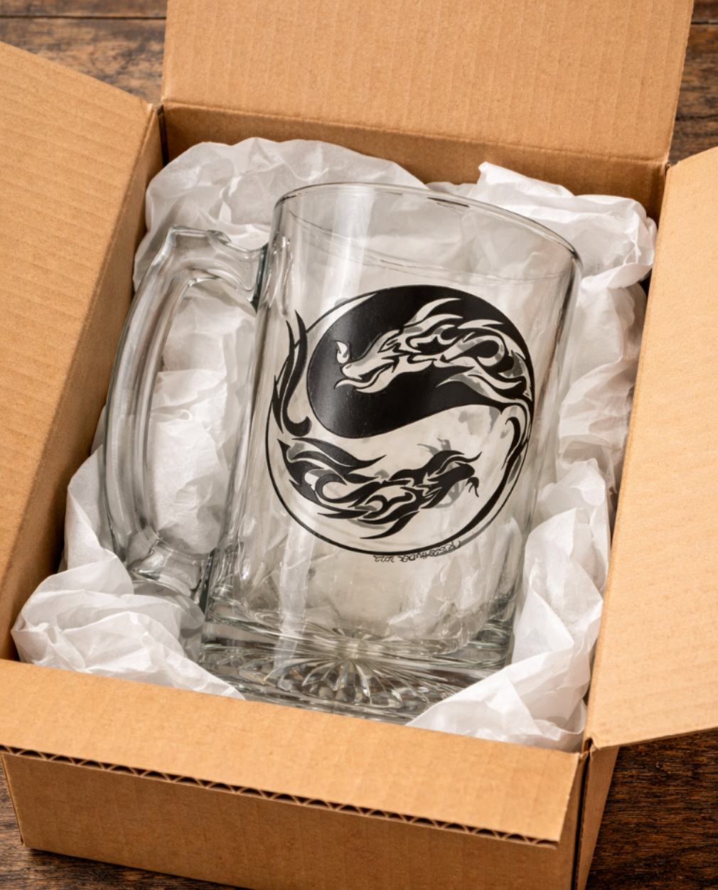 26 oz clear glass beer mug with hand-drawn black yin-yang dragon design, nestled in white tissue paper inside an open brown cardboard shipping box, ready for packaging and delivery.