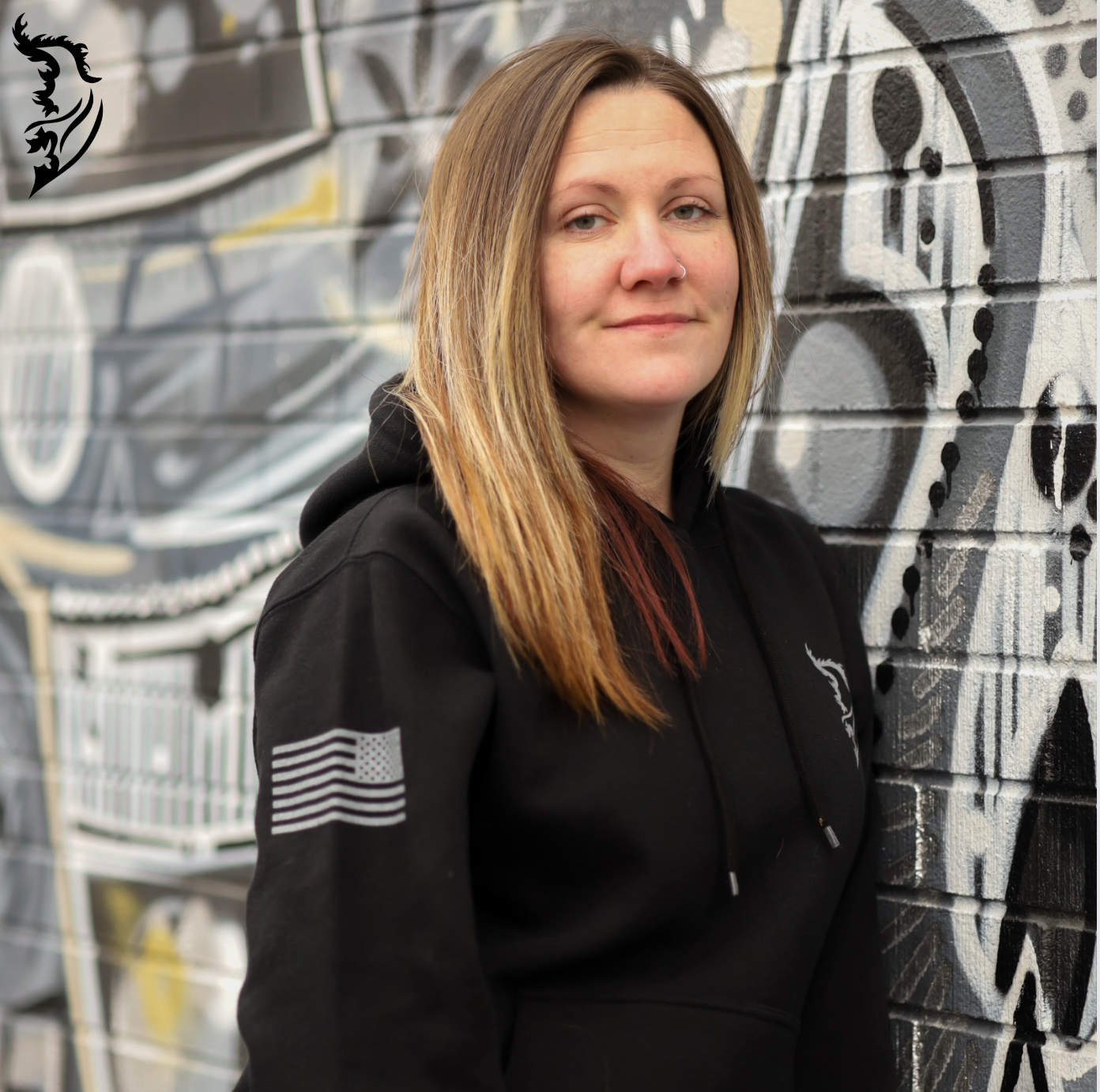 Front view of a black hoodie with a small brand logo on the left chest, featuring a tribute design on the back.