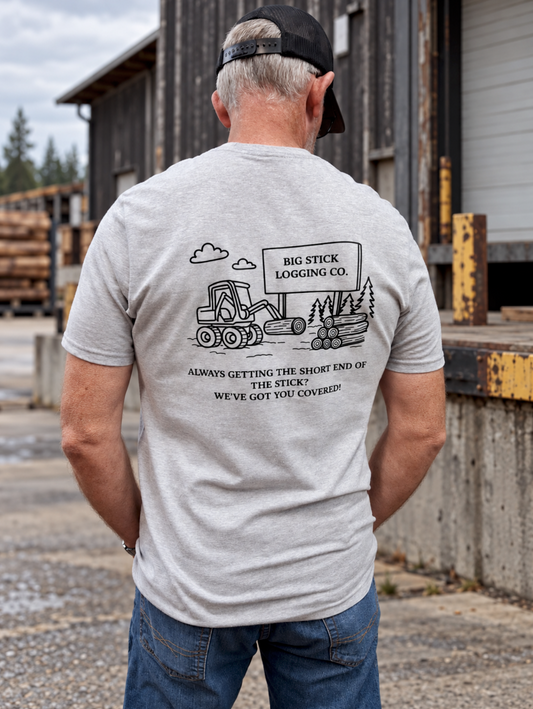 A 6'1" male model wears a heather gray Big Stick Logging Co. T-shirt with an original logging equipment illustration and slogan on the back, blue jeans, and a black cap at an outdoor timber loading dock.