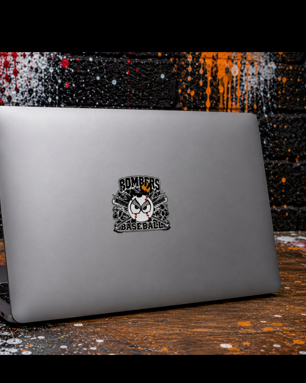 Gray laptop with Bombers Baseball decal sticker centered on the cover, styled against a gritty street-art graffiti wall with splattered paint and urban aesthetic details.