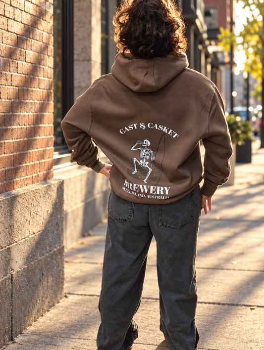 Back view of a brown Cast & Casket Brewery hoodie showing a skeleton holding a drink and fishing rod beneath the words “Cast & Casket,” with “Brewery” below.