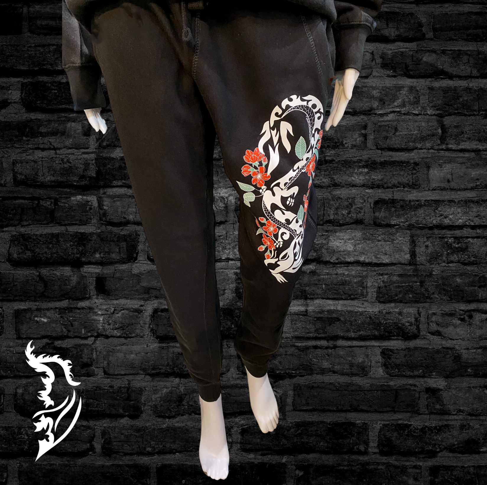 Black joggers featuring a hand-drawn cherry blossom and dragon design down the leg, paired with a white angel wing logo on the back pocket.