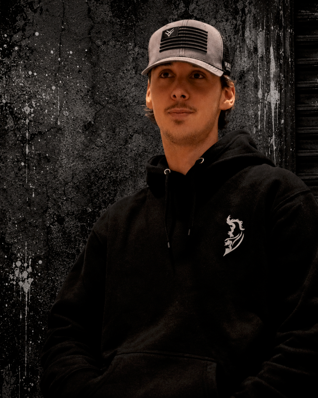 Man wearing black hoodie with white Angel Wing logo on front left chest and gray trucker hat against a dark gritty urban background with industrial textures and moody streetwear-inspired styling by Street Style by Nickola.