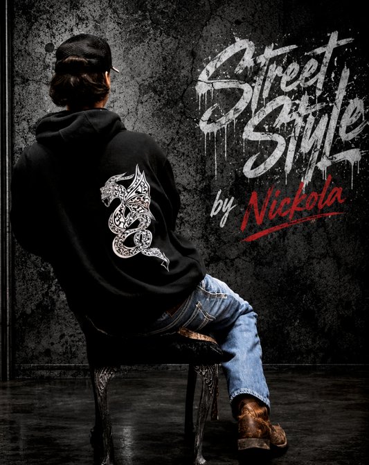 Man wearing black hoodie with hand-drawn dragon by Street Style by Nickola sitting on a vintage chair against a gritty urban streetwear background with bold graffiti-style branding and detailed white dragon artwork on the back of the hoodie.