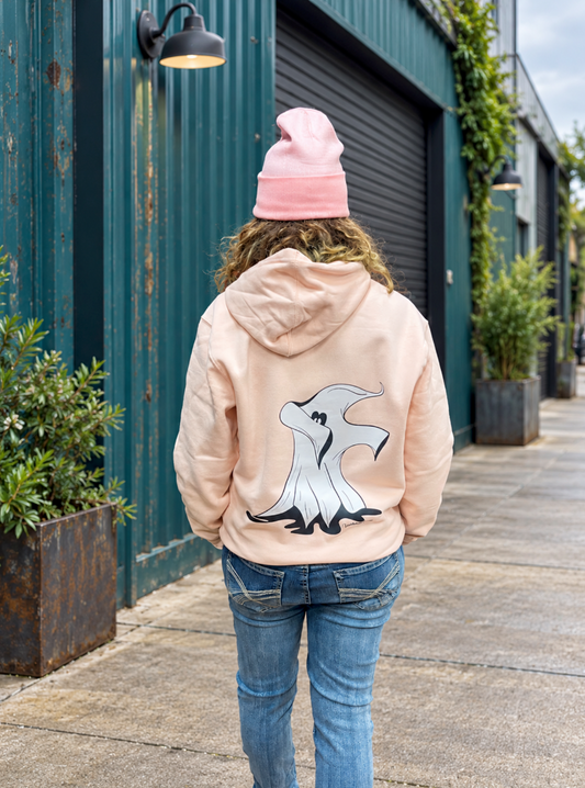 Back view of a woman wearing a pink beanie, pale pink hoodie with a large hand-drawn white-and-black dabbing ghost design, and blue jeans, standing beside a teal industrial building with greenery.