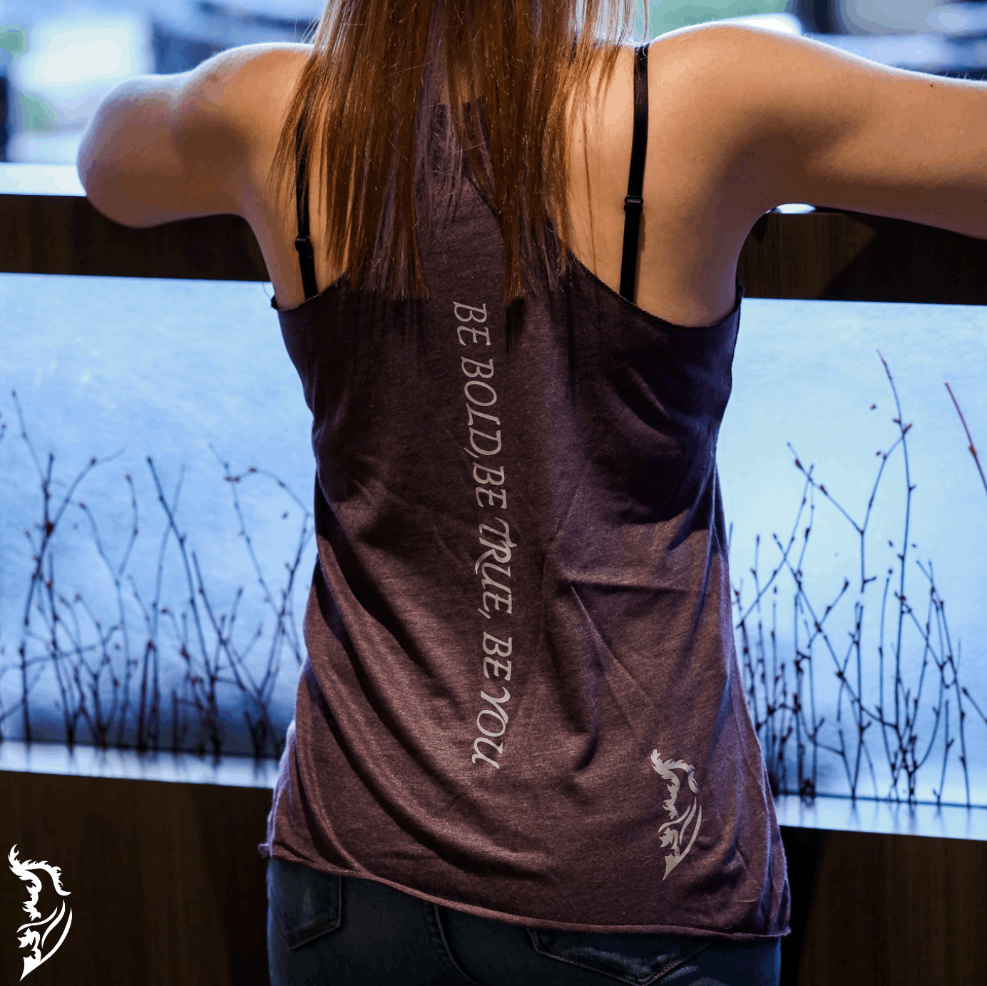 Daisy Racer Back TankBack view of a woman wearing a purple Street Style by Nickola tank top featuring the vertical slogan “Be Bold, Be True, Be You” and Angel Wing logo near the hem, standing indoors by a window railing.