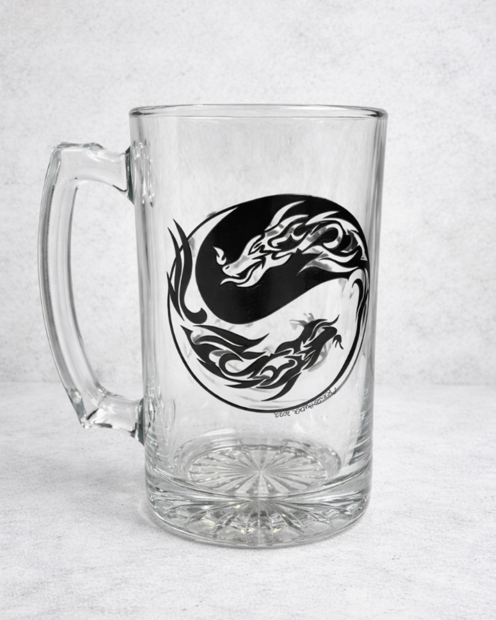 Clear 26 oz glass beer mug featuring a bold hand-drawn black Yin & Yang dragon design, displayed against a light neutral concrete background