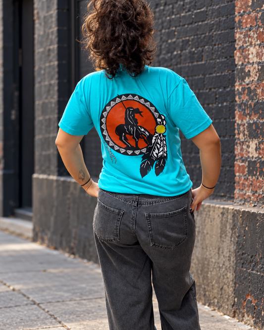 Woman photographed from behind in a Tahiti blue T-shirt and charcoal jeans, standing with her hands on her hips beside a dark brick wall. The shirt has a hand-drawn version of the famous End of the Trail 