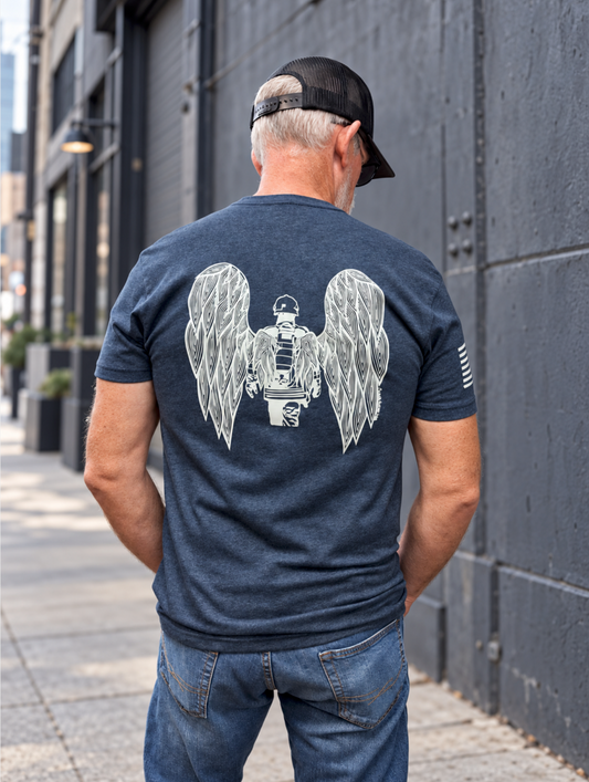 A 6'1" male model wears a dark heather blue T-shirt with a white original design of a fire fighter between two detailed angel wings on the back, paired with blue jeans against a slate-gray street backdrop.