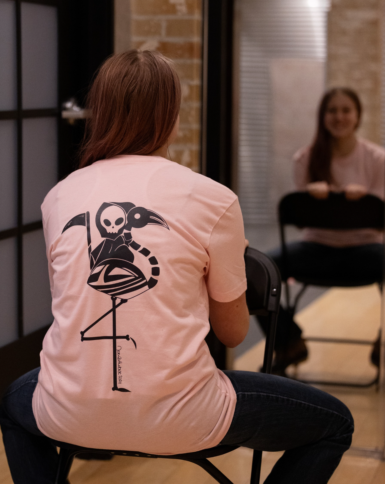 Person wearing a pale pink t-shirt with a hand-drawn grim reaper riding a flamingo  on the back, sitting in a room with a mirror.