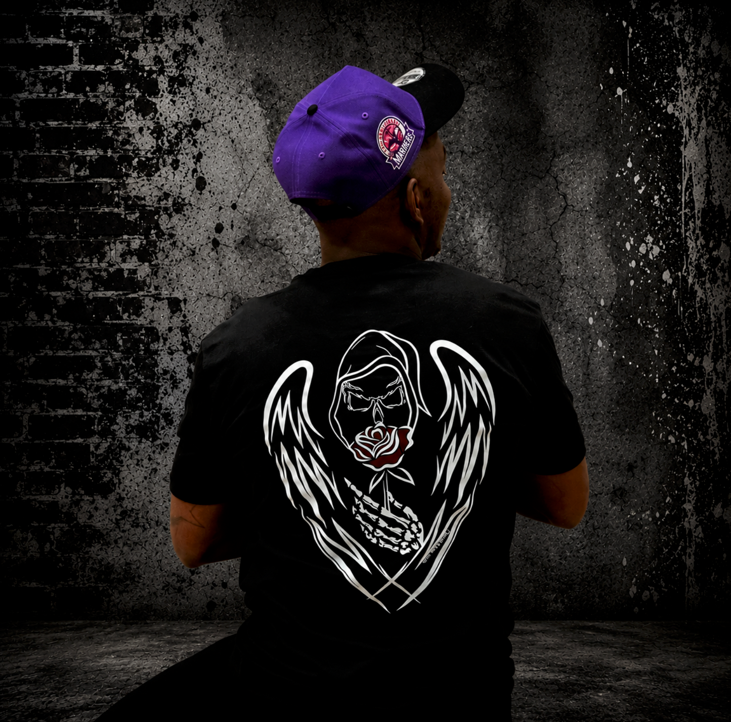 Man wearing black Grim Reaper graphic t-shirt with hand-drawn grim reaper with white skeletal angel wings and red rose artwork on the back, paired with a purple Mariners cap against a gritty urban-style background.
