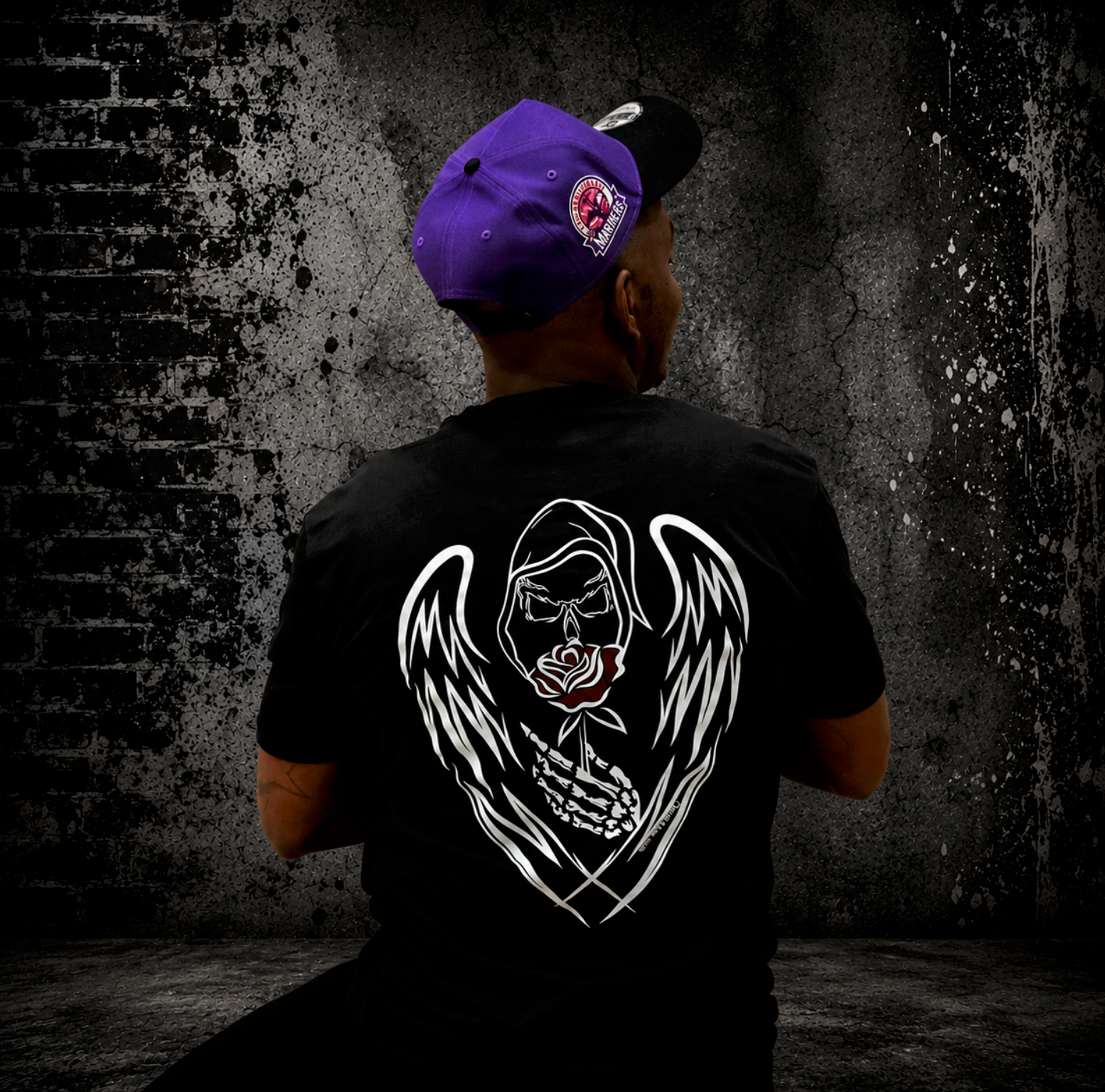 Man wearing black Grim Reaper graphic t-shirt with hand-drawn grim reaper with white skeletal angel wings and red rose artwork on the back, paired with a purple Mariners cap against a gritty urban-style background.