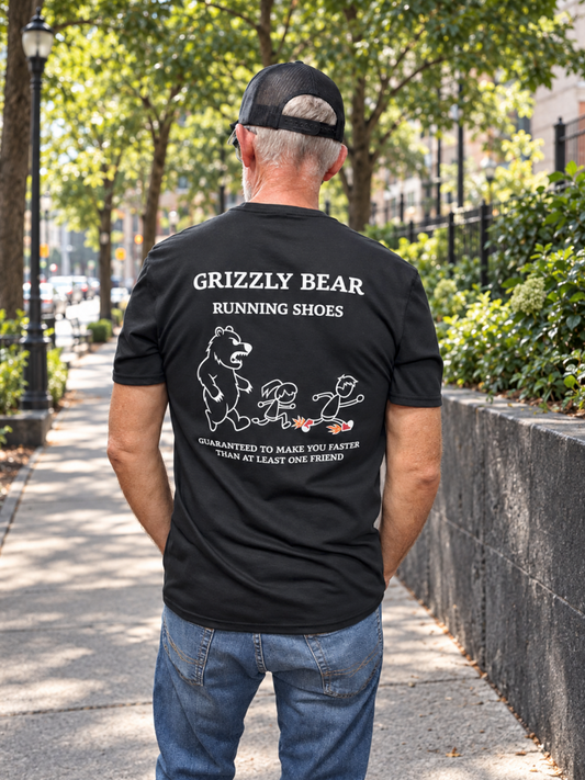 Back view of a male model in a black Grizzly Bear Running Shoes T-shirt on a tree-lined sidewalk. The large back print shows a bear chasing two runners and the words “Guaranteed to make you faster than at least one friend.”