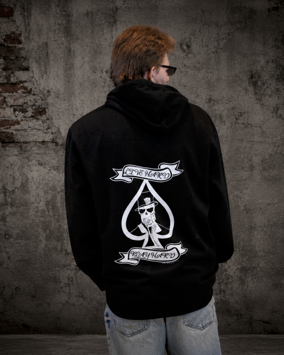 Man modeling black “Live Hard Play Hard” hoodie by Street Style by Nickola standing in front of an industrial concrete wall 