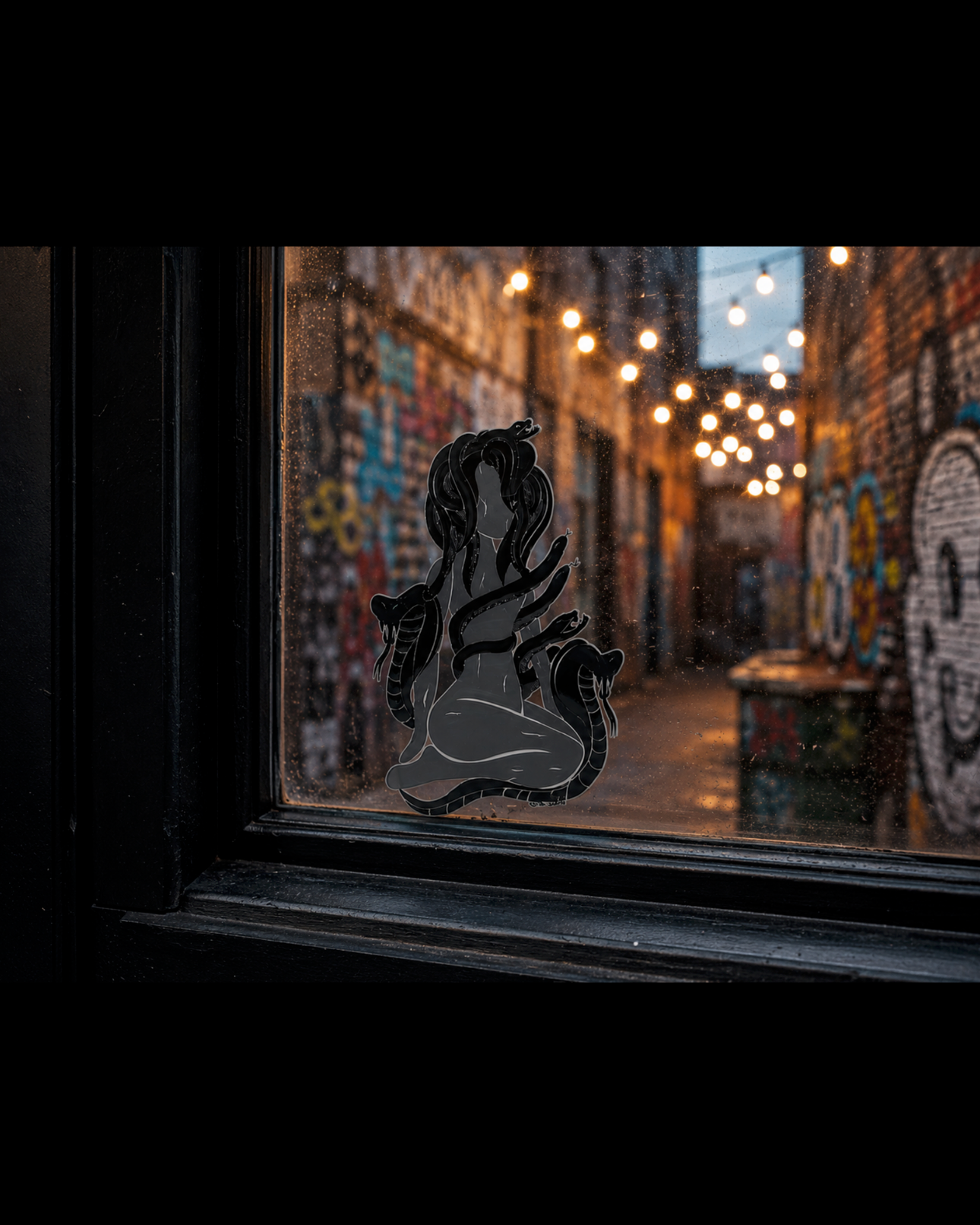 Black Hand-drawn Medusa UV sticker decal displayed on a window overlooking a graffiti-covered alley with glowing string lights