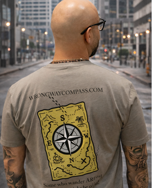 Back view of a man wearing a tan Wrong Way Compass graphic T-shirt featuring a map and compass design, standing in an urban city street setting with buildings and lights in the background.