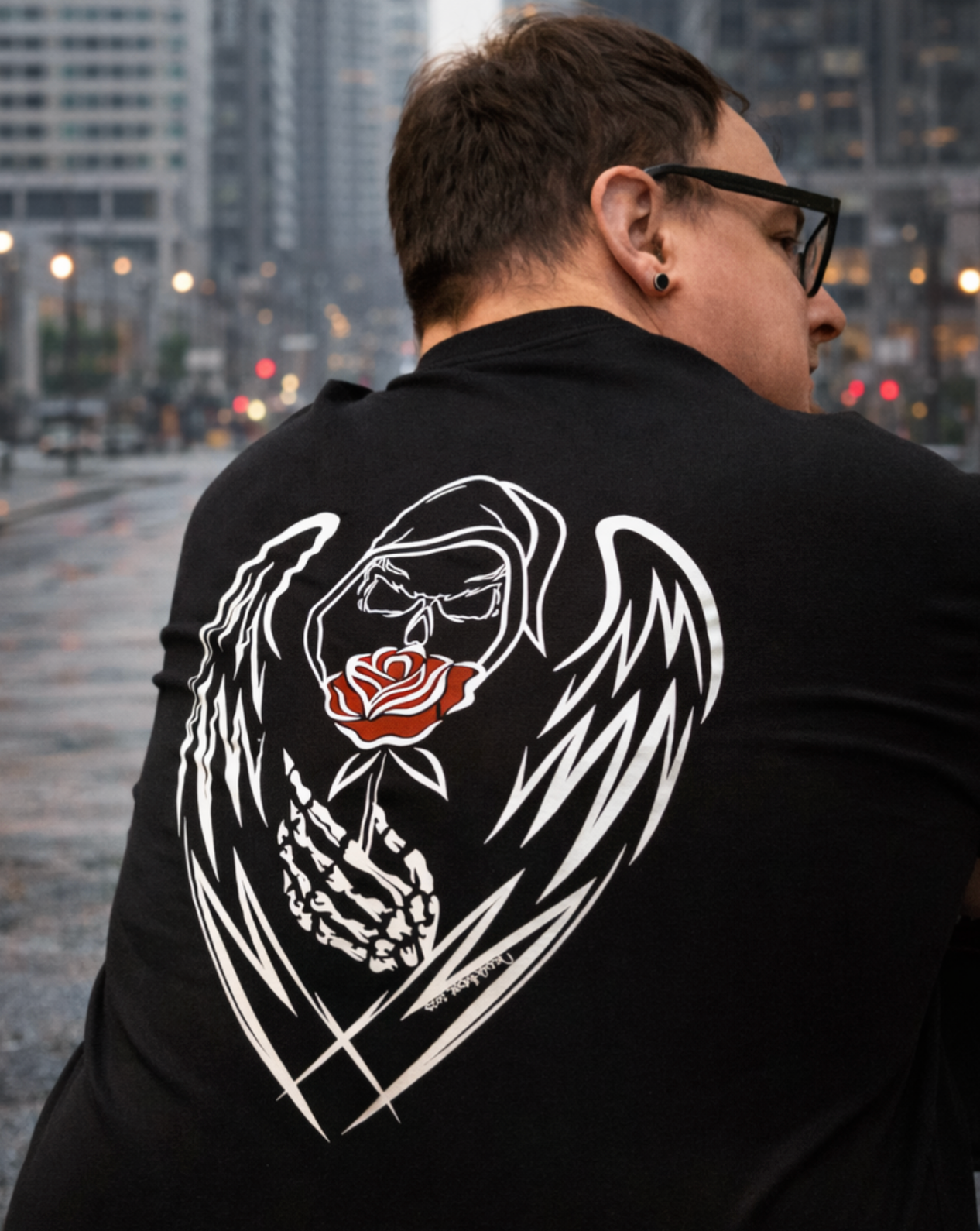 Man wearing a black Street Style by Nickola T-shirt with hand-drawn Grim Reaper with a Rose  standing on a rainy urban city street with buildings and glowing streetlights in the background.