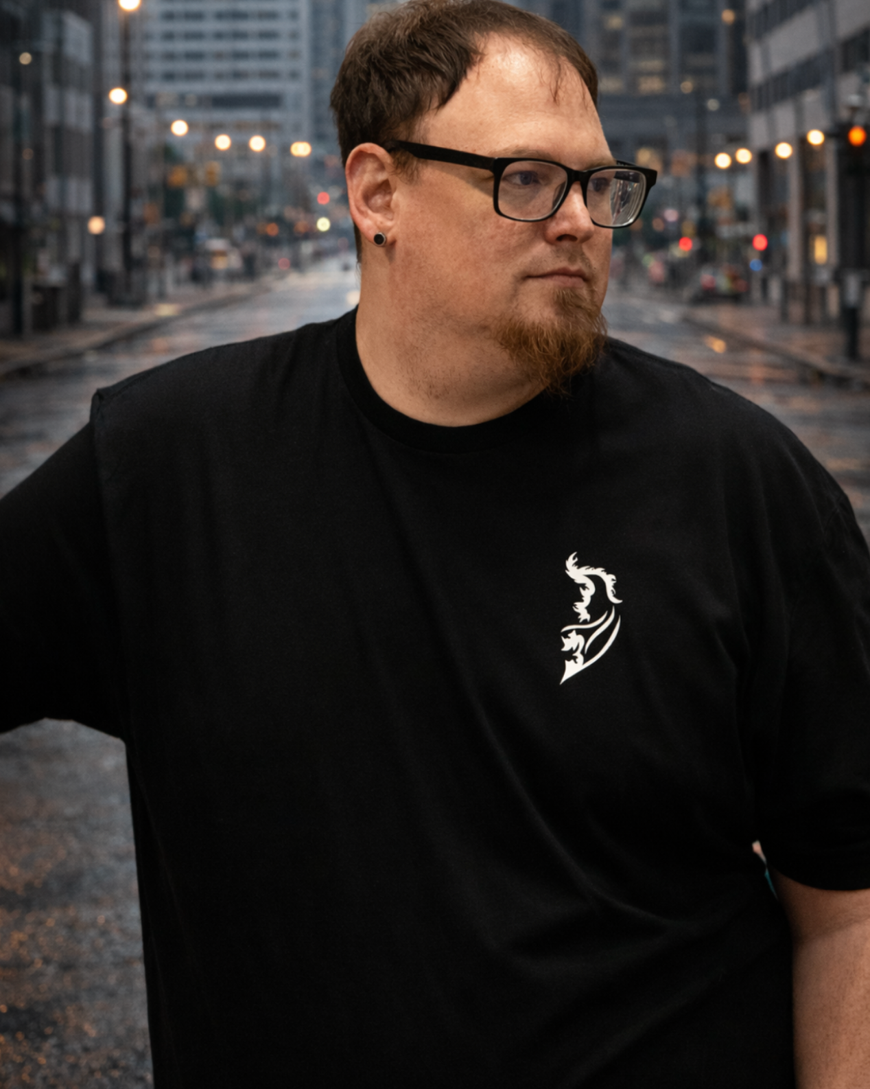 Man wearing a black Street Style by Nickola T-shirt with Angel Wing Logo on left chest, standing on a wet urban city street with buildings and streetlights in the background.