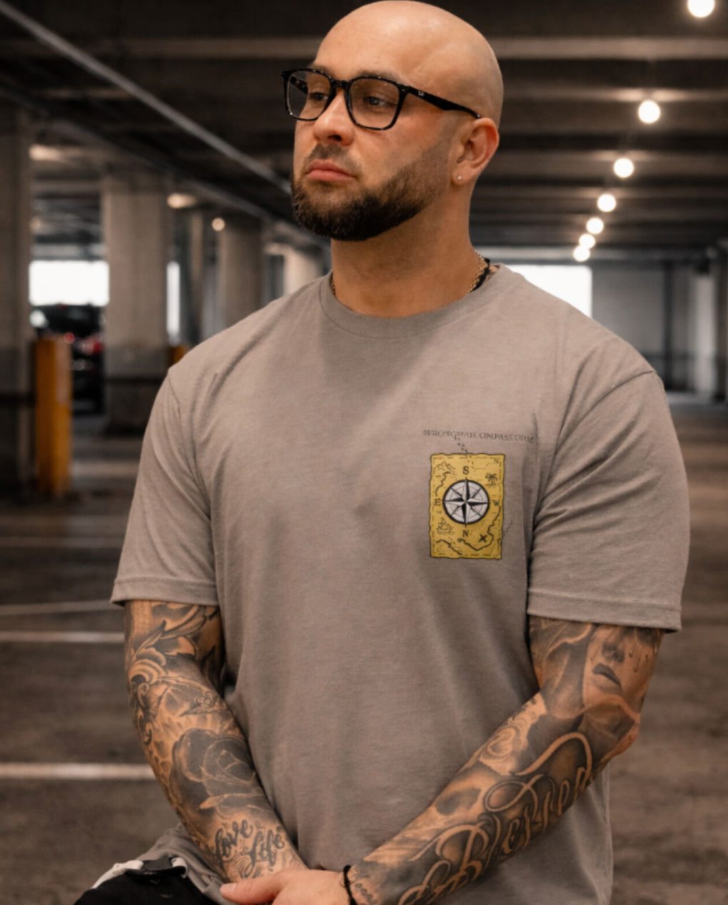 Man wearing a Warm Grey Street Style by Nickola Wrong Way Compass.Com graphic T-shirt standing in an urban parking garage setting, showing tattooed arms and casual streetwear style.