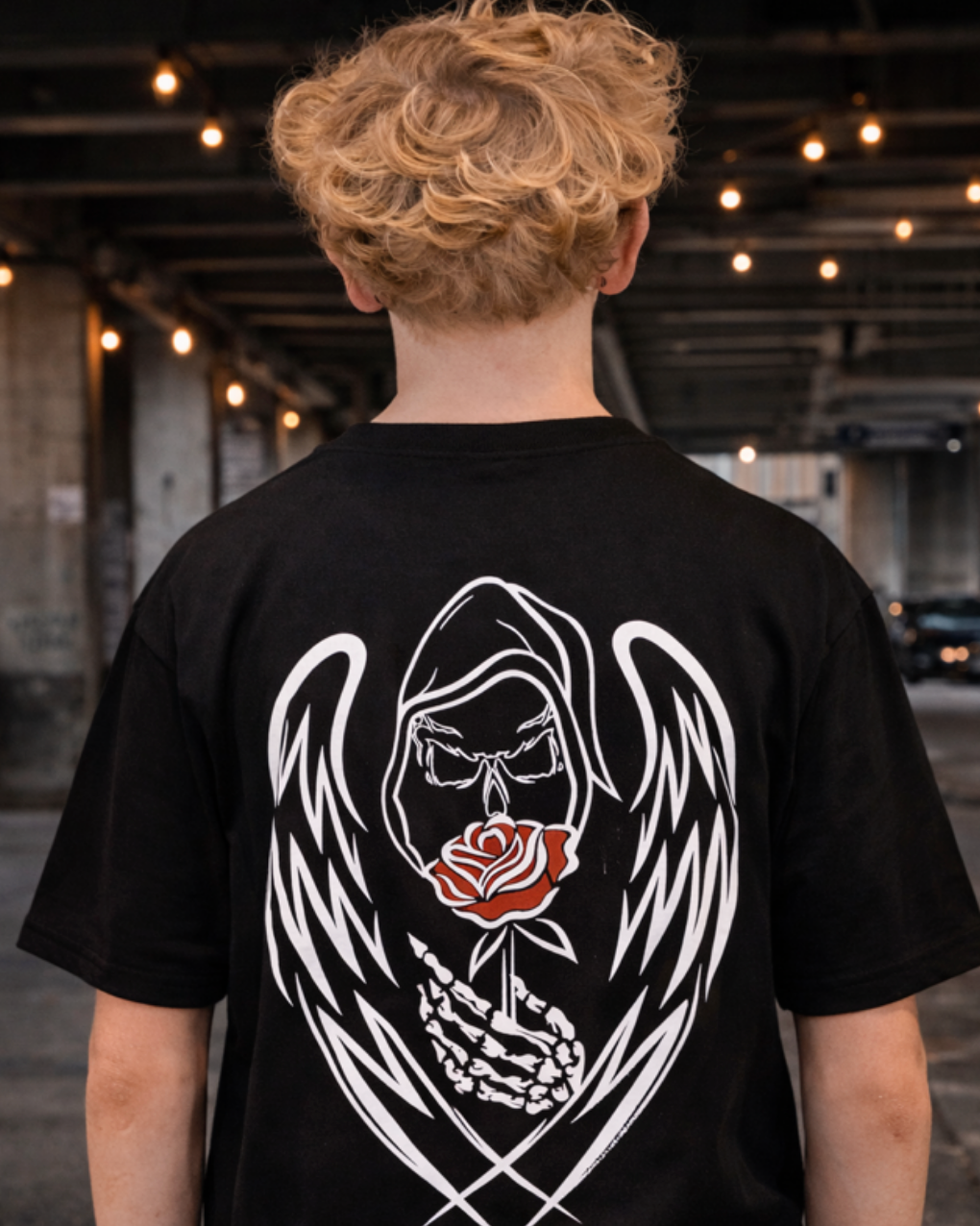 Back view of a model wearing a black Street Style by Nickola Grim Reaper Rose T-shirt featuring a hand-drawn white winged Grim Reaper holding a red rose, photographed in an urban underpass setting with warm hanging lights.
