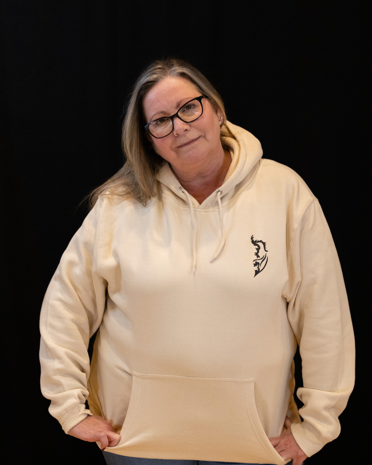 Woman wearing a Sandstone Street Style by Nickola hoodie with a black Angel Wing logo on front left chest,, standing against a dark background.