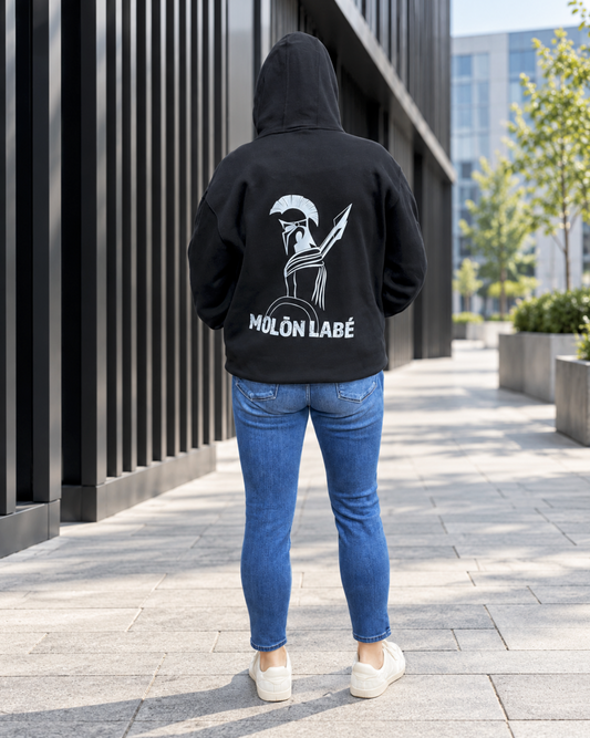 Back view of a model wearing a black hoodie with a hand-drawn white Spartan warrior design and “MOLŌN LABÉ” lettering, blue jeans, and cream sneakers beside a modern black metal building.