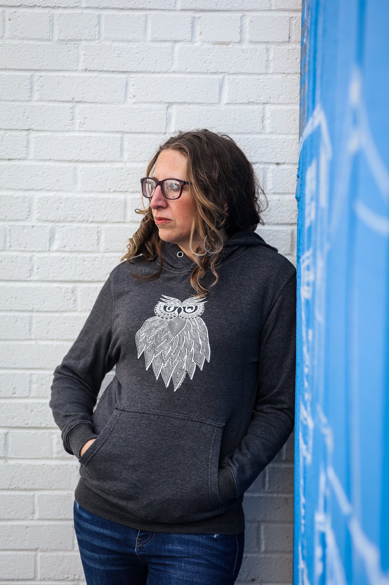 Female Model wearing Owl Hoodie leaning against brick wall with hand-drawn lined owl on front 