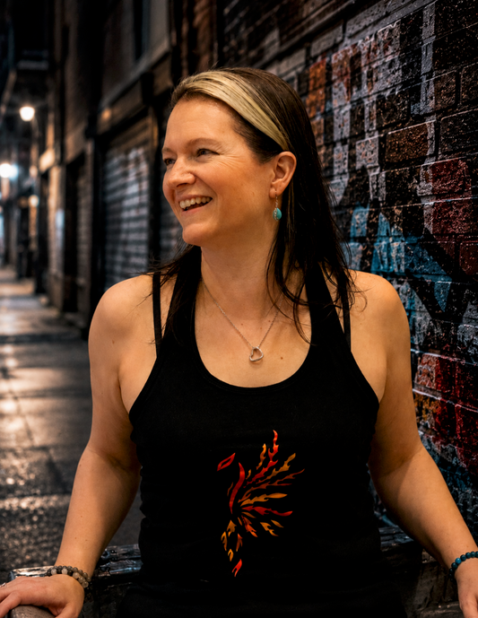A smiling woman models a black racerback tank top featuring an original red and orange phoenix on the front. She stands in a moody urban alley with graffiti-covered brick walls and glowing streetlights.