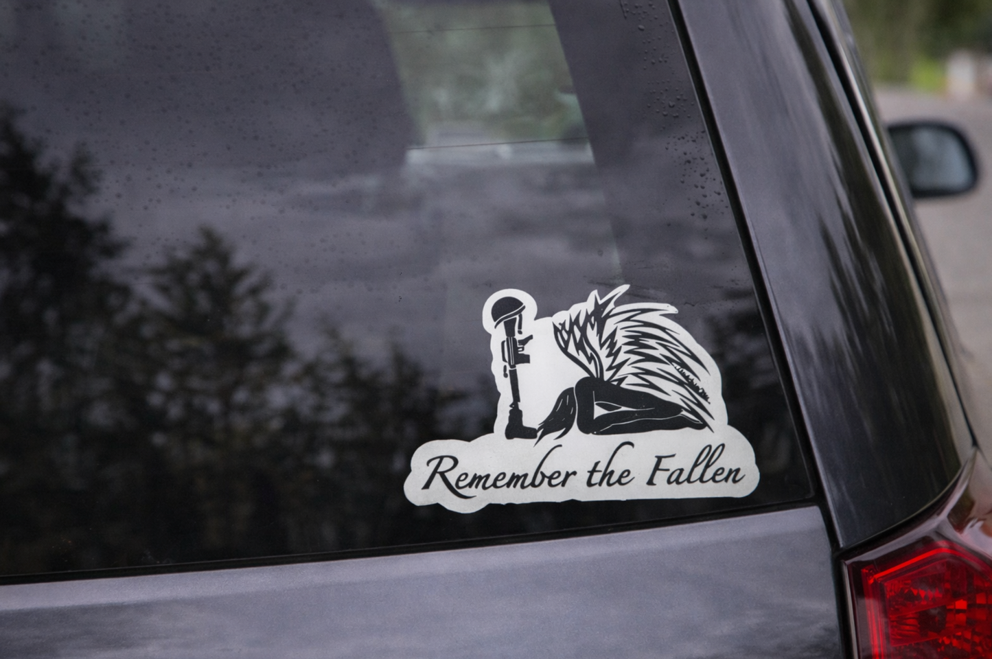 Black-and-white “Remember the Fallen” memorial sticker displayed on the lower right corner of a dark blue car’s rear window. The design features a kneeling angel with detailed wings beside a rifle topped with a helmet, with the words “Remember the Fallen” written beneath. The car’s glossy surface reflects trees and sky in the background.