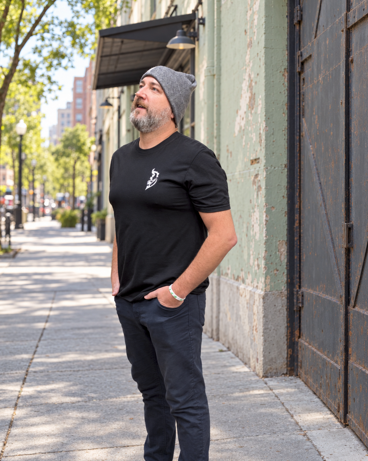 Bearded man wearing a gray beanie, black T-shirt with a small white Angel Wing logo and dark pants, looking upward with his hands in his pockets beside a weathered sage-green warehouse on a tree-lined sidewalk.
