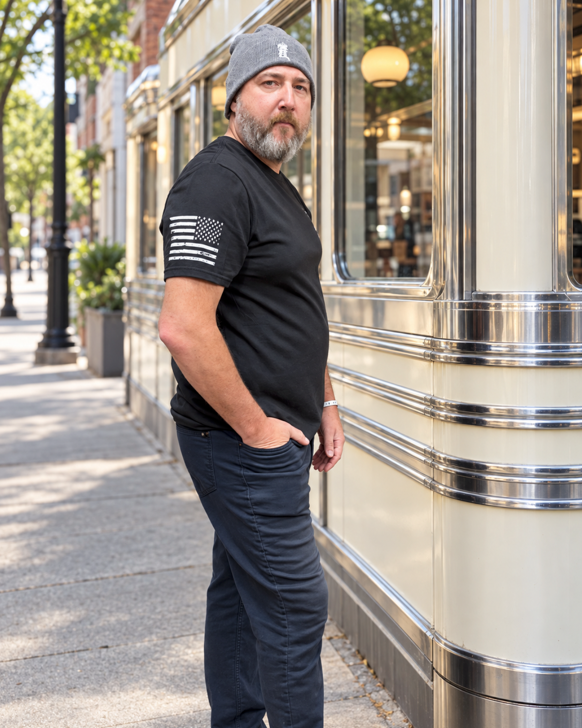 Bearded man wearing a gray beanie, black T-shirt with a distressed white American flag on the sleeve, and dark pants, standing with one hand in his pocket beside a cream-and-chrome diner.