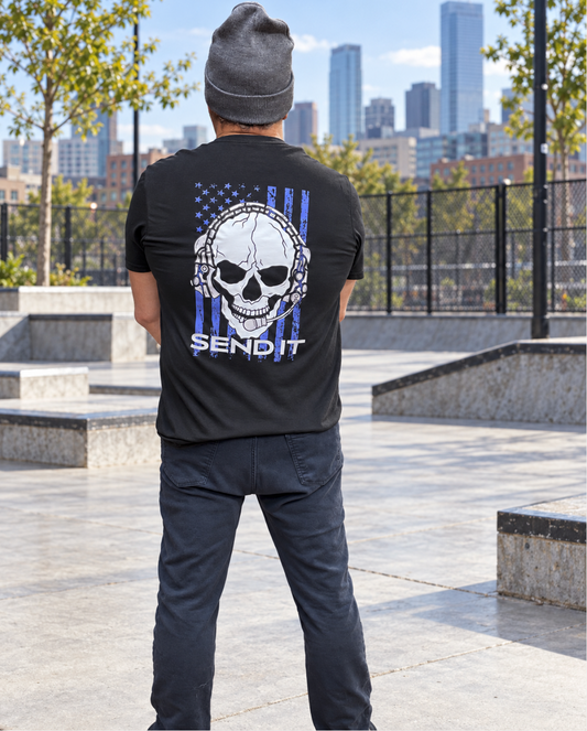 Back view of a man wearing a gray beanie and black T-shirt featuring a white skull with a headset over a distressed blue-purple American flag and “SEND IT” text, standing in an urban skate plaza.