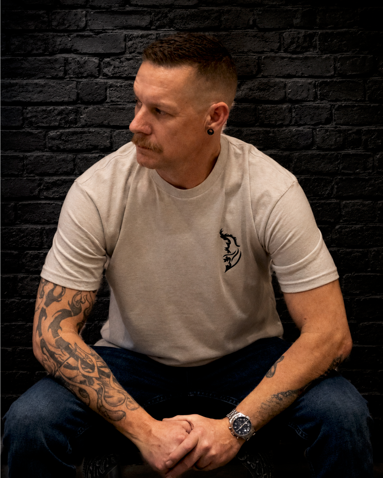 Man wearing a silk graphic t-shirt with a black Angel Wing logo, seated against a dark black brick wall background. The model features tattooed arms, dark jeans, and a moody streetwear-inspired aesthetic by Street Style by Nickola.