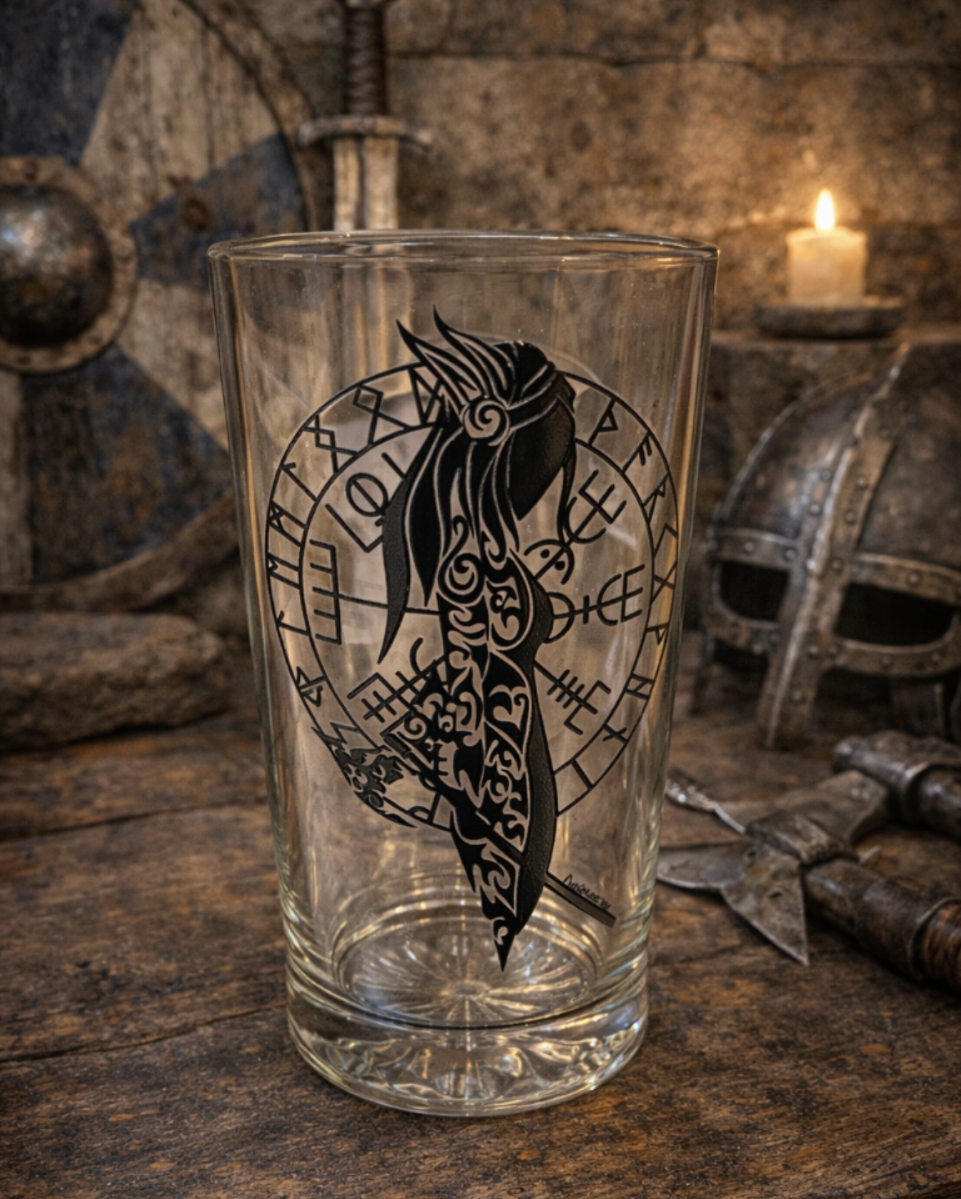 Clear pint glass featuring a hand-drawn bold black Norse shield maiden silhouette with rune circle design, displayed on a rustic wooden table with Viking helmet, shield, sword, and candlelit background.
