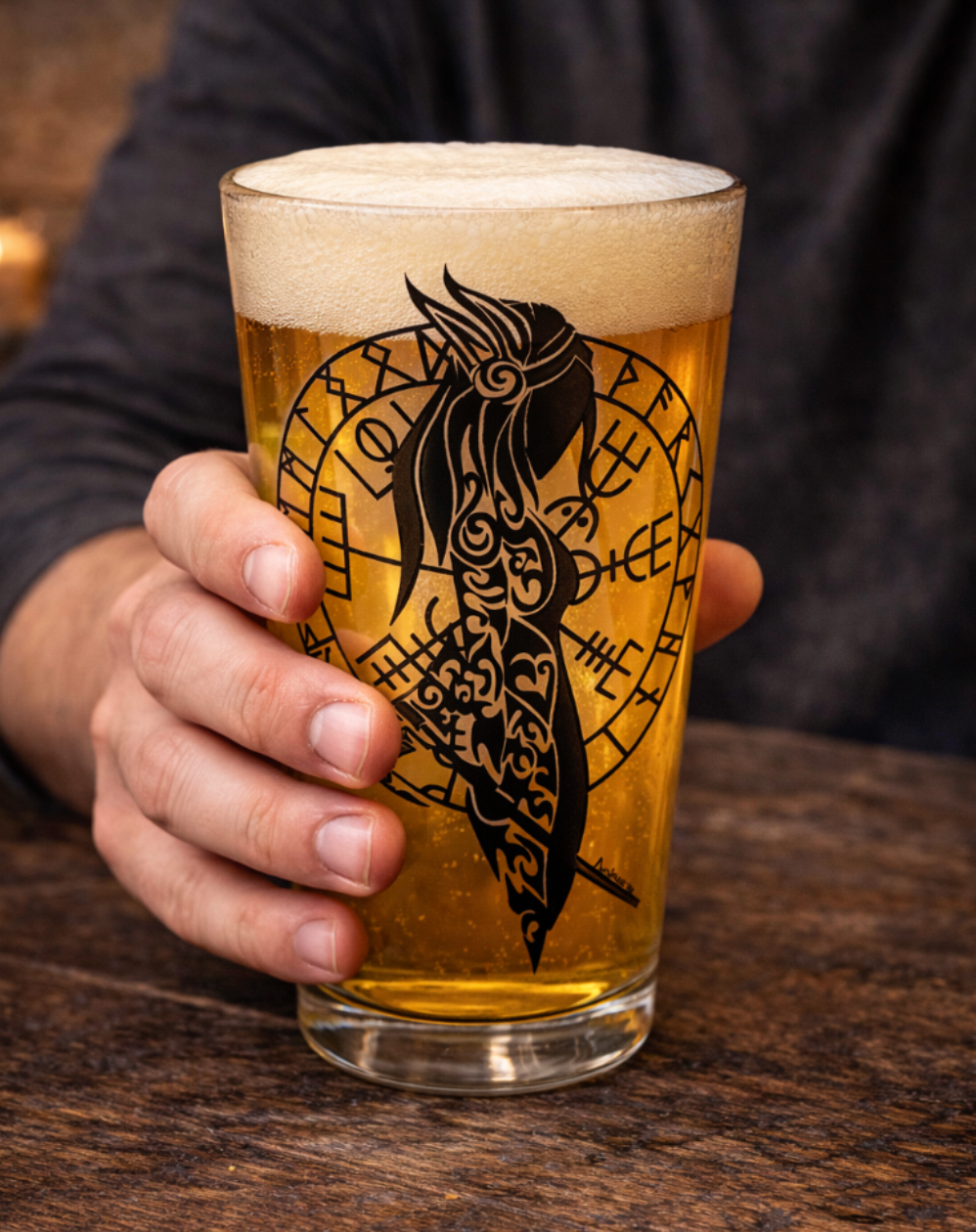 Pint glass filled with golden beer and thick foam, featuring a hand-drawn bold black Norse shield maiden silhouette over a rune circle design, set on a rustic wooden table with Viking helmet, sword, candle, and shields in the background.