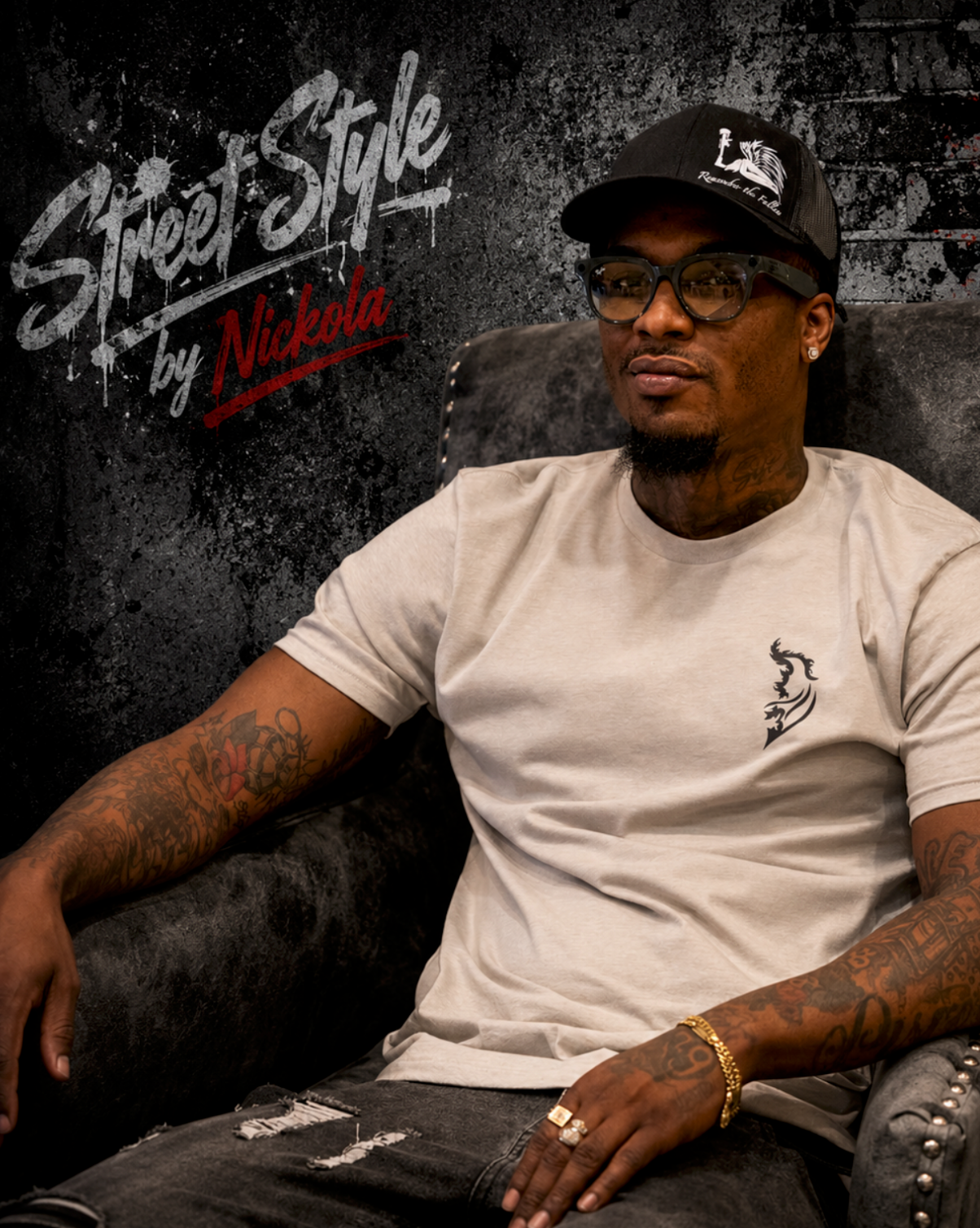 Man wearing Silk Street Style by Nickola t-shirt, black hat, and glasses sitting in a chair against a gritty urban graffiti-style background with dark industrial textures and streetwear-inspired branding.