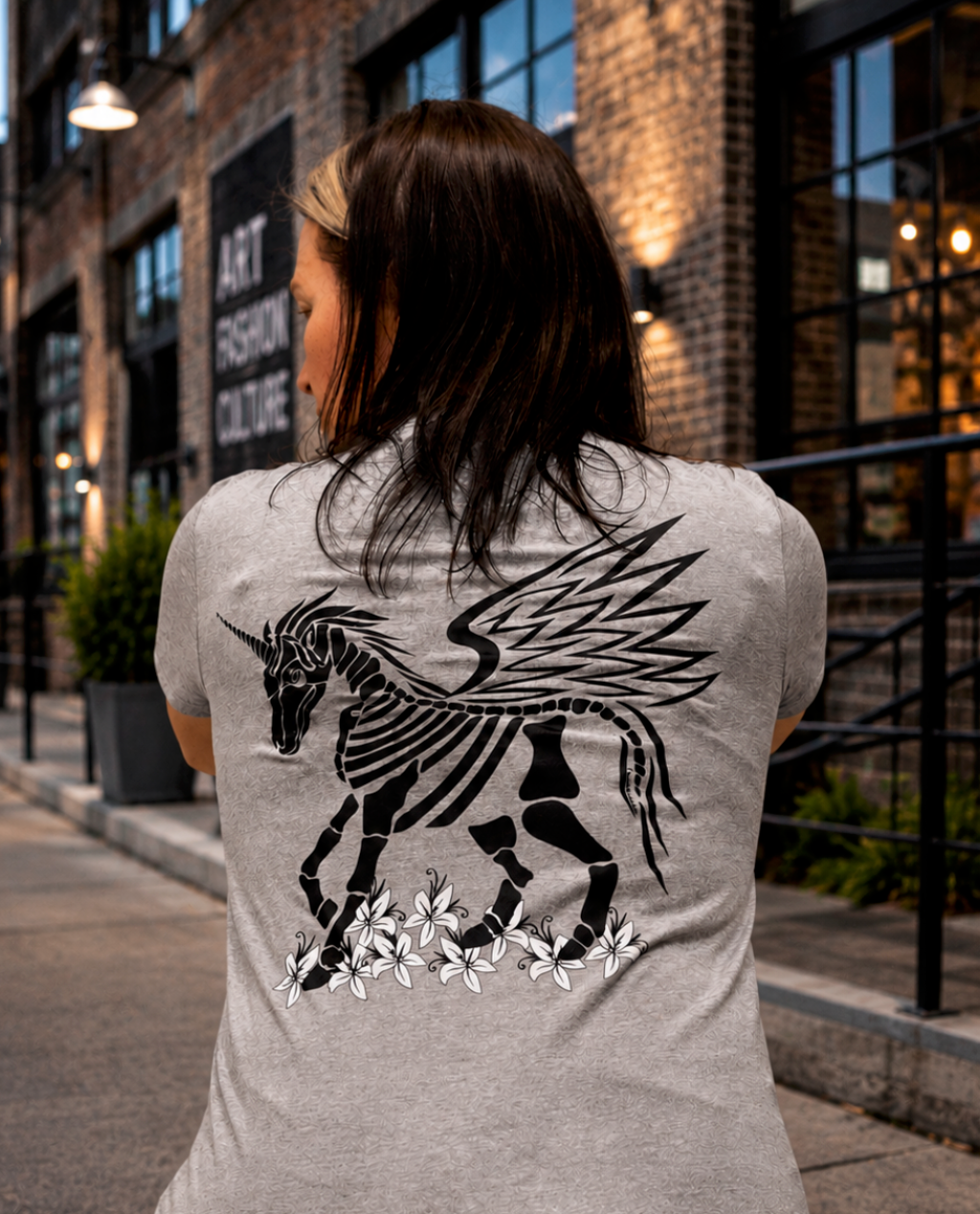 Back view of a person wearing a heather gray T-shirt featuring an original hand-drawn winged skeletal unicorn graphic in black and white with floral accents along the bottom. The person stands in front of a modern urban brick building with large industrial windows, warm exterior lighting, and a clean city sidewalk.