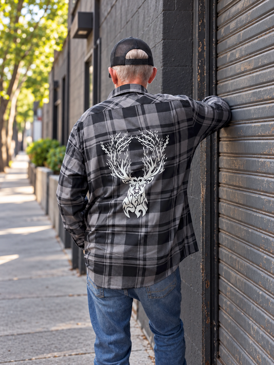 Back view of a male model wearing a gray and black plaid flannel with a large hand-drawn white stag design across the back, standing beside a dark storefront shutter.