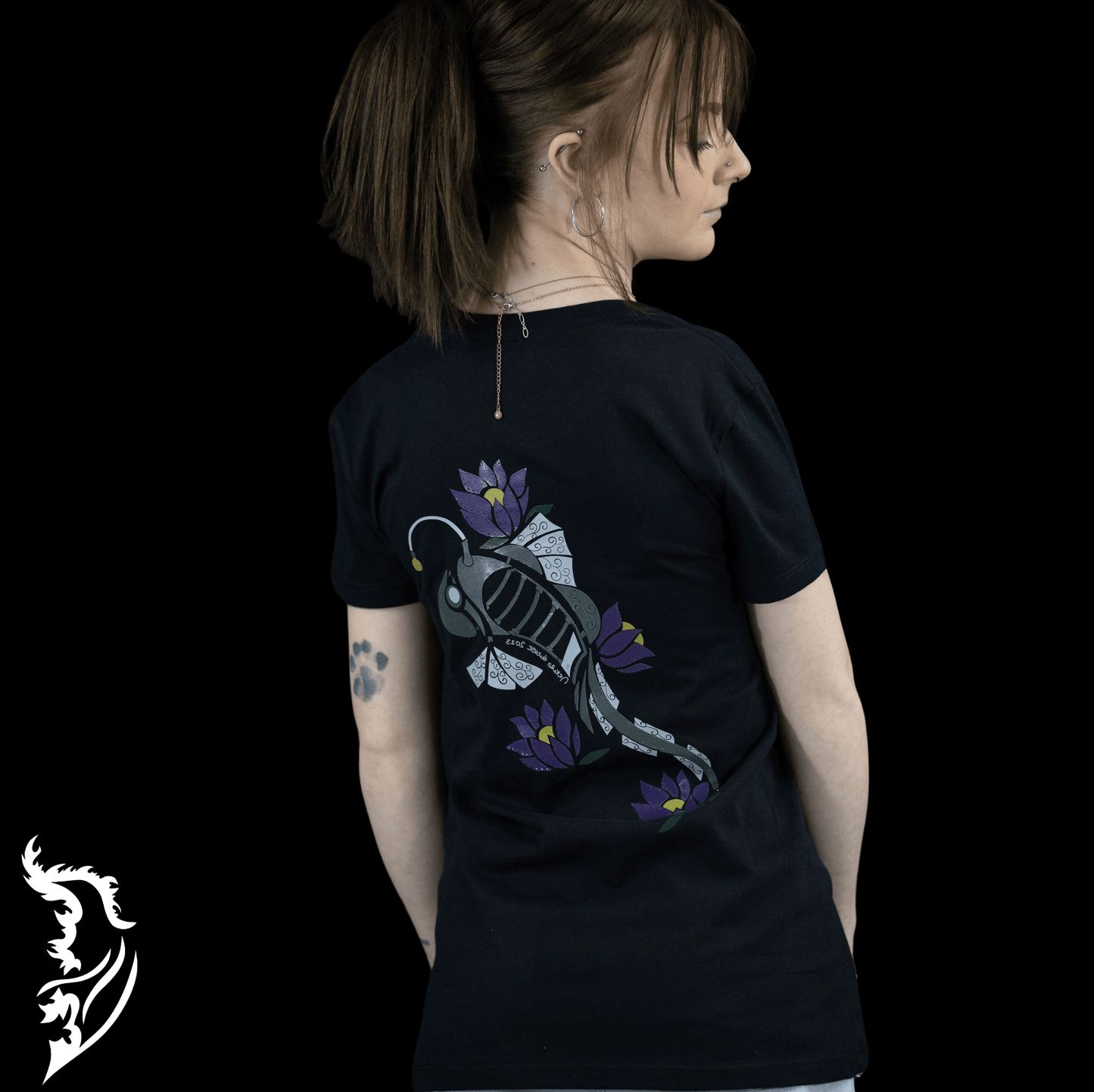 Woman wearing a black Street Style by Nickola slim-fit T-shirt featuring a hand-drawn steampunk fish design with purple lotus flowers on the back, photographed against a dark background.