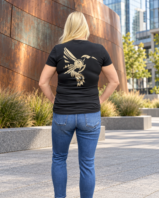 Back view of a blonde woman wearing a fitted black T-shirt with a large hand-drawn bronze Steampunk Hummingbird and blue jeans, standing with her hands on her hips beside a curved copper wall in a city plaza.