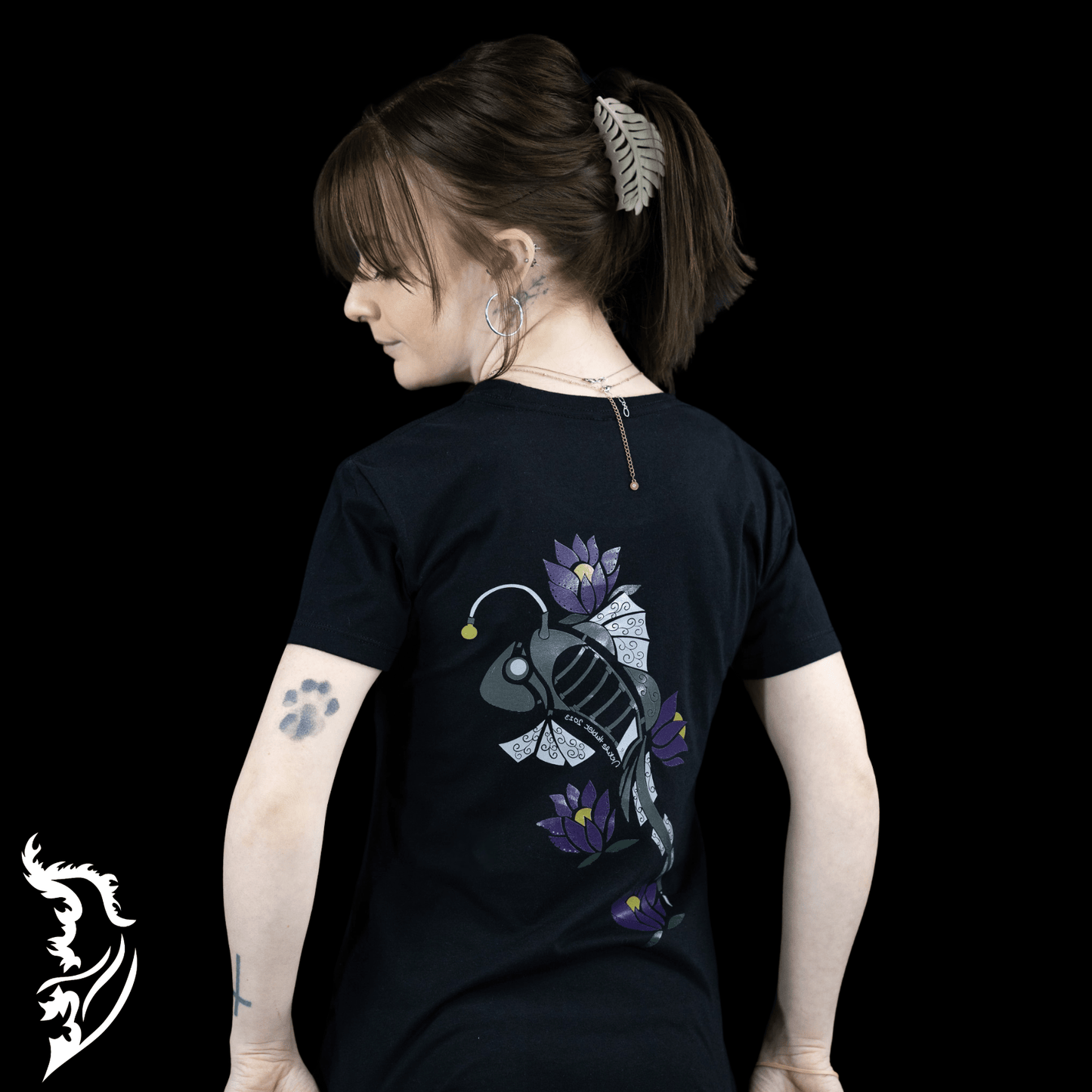 Woman wearing a black Street Style by Nickola slim-fit T-shirt featuring a hand-drawn steampunk fish design with purple lotus flowers on the back, photographed against a dark background.