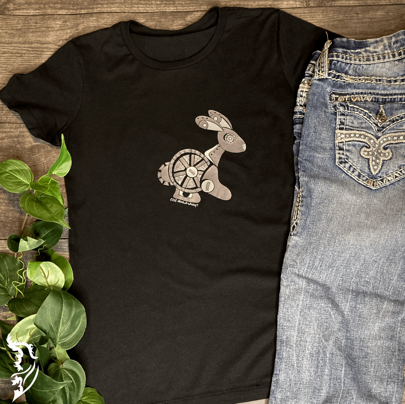 Flat lay of a black Street Style by Nickola T-shirt featuring a hand-drawn steampunk rabbit design, styled with light blue embroidered jeans and green leafy plant accents on a wooden background.