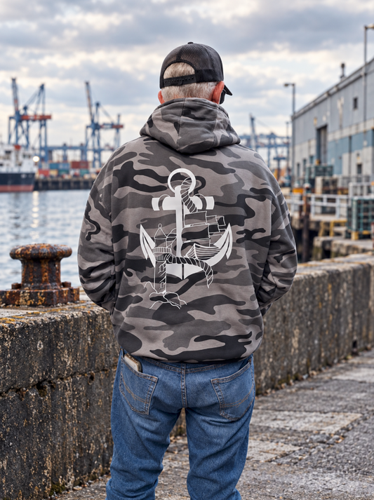 Back view of a male model wearing a gray and black camouflage hoodie with a large hand-drawn white anchor, lighthouse, and sailing ship design, standing beside an industrial harbor.