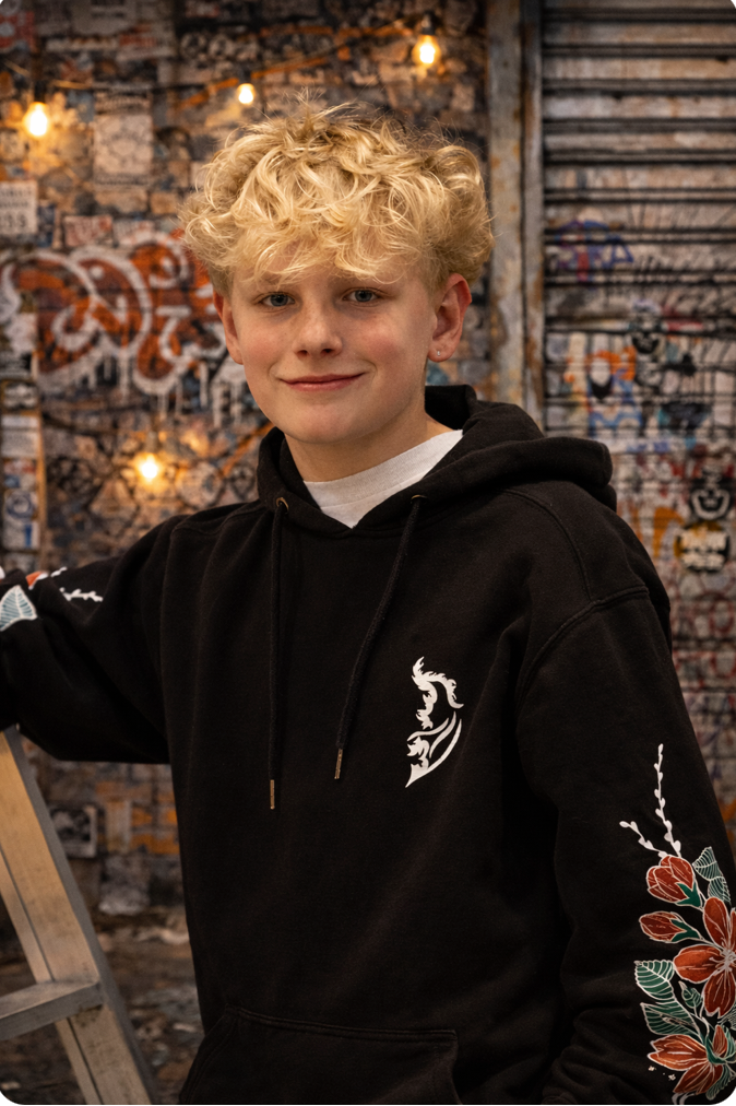 Teen model wearing a black Street Style by Nickola hoodie with Angel Wing Logo on front left chest and cherry blossom  design down sleeves, posing in front of an urban graffiti wall with warm string lights during a community photo shoot.
