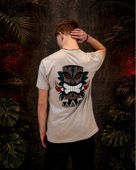 Back view of a model wearing a silk Tiki men’s T-shirt with a large hand-drawn mask graphic in black, green, red, and white, posed against a dark wall framed by tropical leaves.