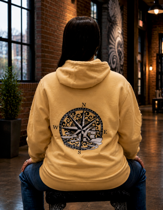 Back view of a person seated on a decorative stool wearing a soft yellow hoodie featuring an original hand-drawn nautical compass and sailing ship design across the back. The hoodie is showcased in a warm industrial loft with exposed brick walls, large black-framed windows, polished concrete floors, and ambient lighting.