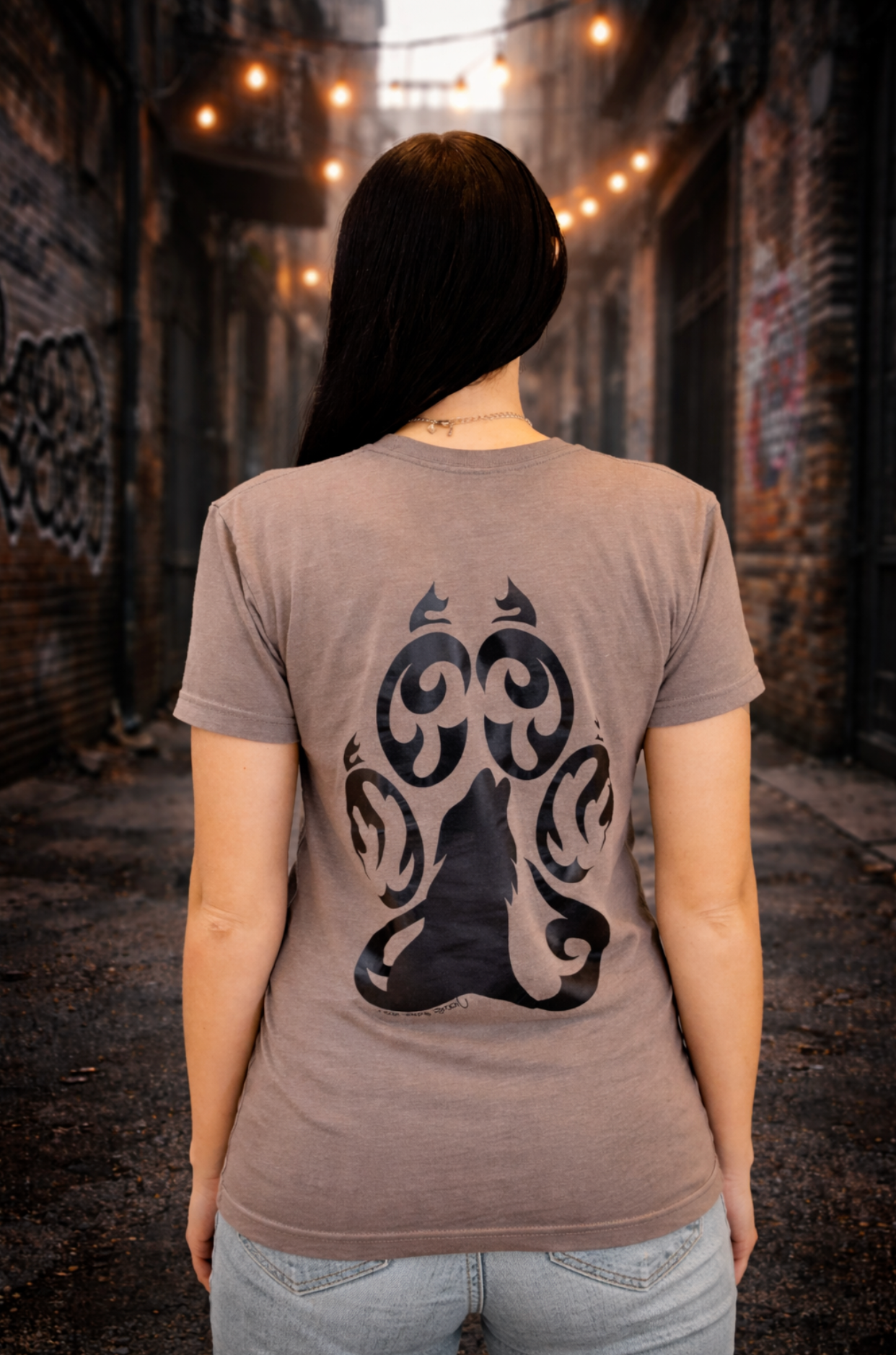 Woman modeling gray wolf design t-shirt from Street Style by Nickola in an urban alley background with graffiti and warm string lights, showcasing bold back print artwork.