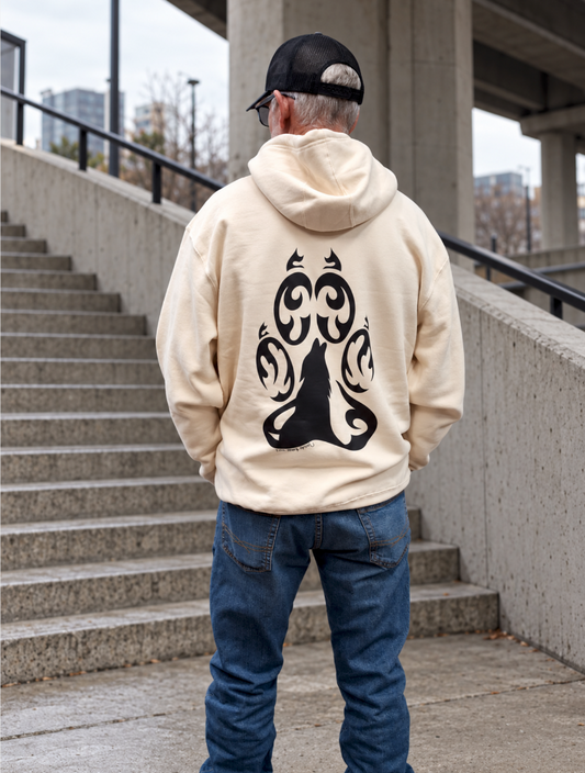 A 6'1" male model wears an XL sandstone hoodie with a large hand-drawn black tribal bear paw design, blue jeans, and a black cap beside an outdoor concrete stairway.