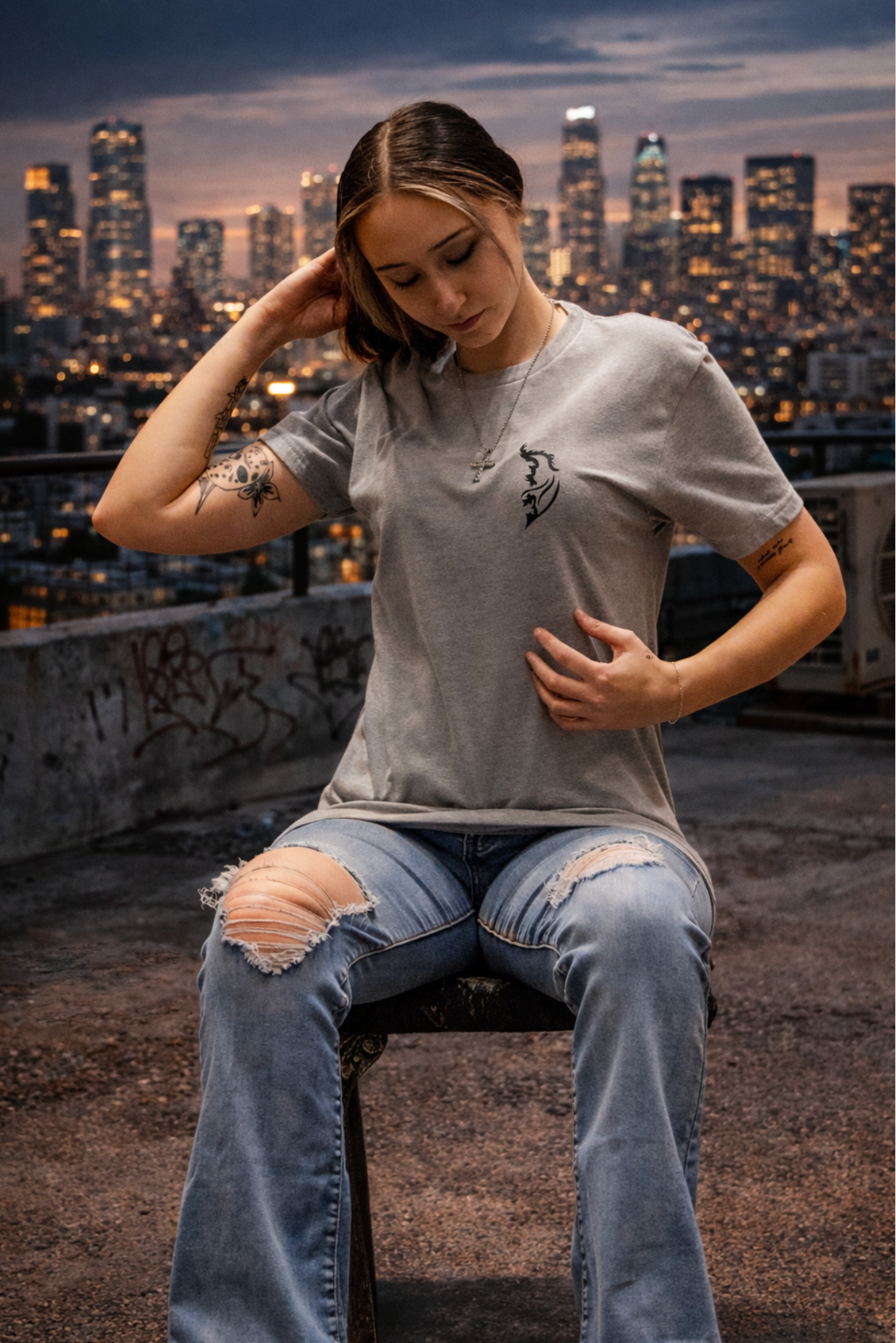 Female model wearing silk Street Style by Nickola t-shirt with Black Angel Wing logo, seated on a rooftop overlooking a city skyline at sunset, styled in urban streetwear setting.